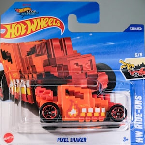 Puede incluir: Un coche de juguete fundido a presión Hot Wheels Pixel Shaker naranja y rojo en su embalaje original. El coche presenta un diseño pixelado y ruedas negras con llantas rojas. El embalaje incluye el logotipo de Hot Wheels y el texto "Pixel Shaker".