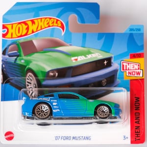 Könnte beinhalten: Ein blaues und grünes Hot Wheels Spielzeugauto Ford Mustang 2007 mit Falken Reifen. Das Auto befindet sich auf einem blauen und weißen Hintergrund mit den Worten "Then and Now" und "Hot Wheels" auf der Verpackung.