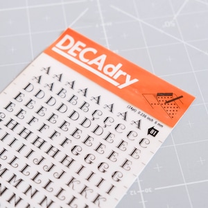 May include: A sheet of black letter stickers on a white background. The letters are in a decorative font and include A through N. The sheet is labeled "DECAdry" and includes the size of the letters in inches and millimeters.