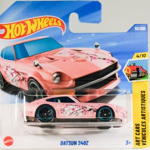 May include: Pink Datsun 240Z die-cast toy car with cherry blossom graphics. The car has black wheels with blue rims and is packaged in a Hot Wheels blister pack. The package includes the text "Art Cars" and "Datsun 240Z".