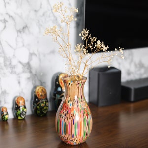 May include: A colorful vase made from pencils, with a bouquet of white flowers. The vase is a unique and decorative piece for any home.