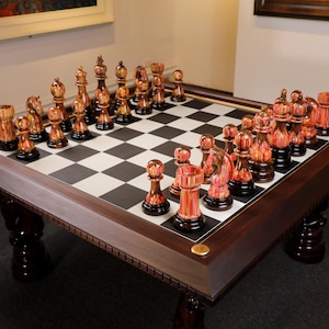 May include: A large, square chess table with a dark wood frame and legs. The chessboard has a black and white checkered pattern. The chess pieces are a mix of orange and gold tones. The table is set up for a game of chess.