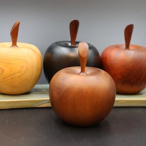 May include: Four wooden apples, each carved from a different type of wood, with natural wood grain patterns. The apples are arranged on a green and brown ceramic tile.