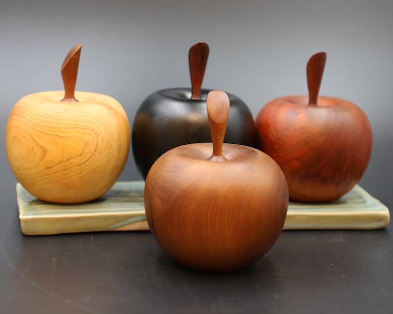 May include: Four wooden apples, each carved from a different type of wood, with natural wood grain patterns. The apples are arranged on a green and brown ceramic tile.