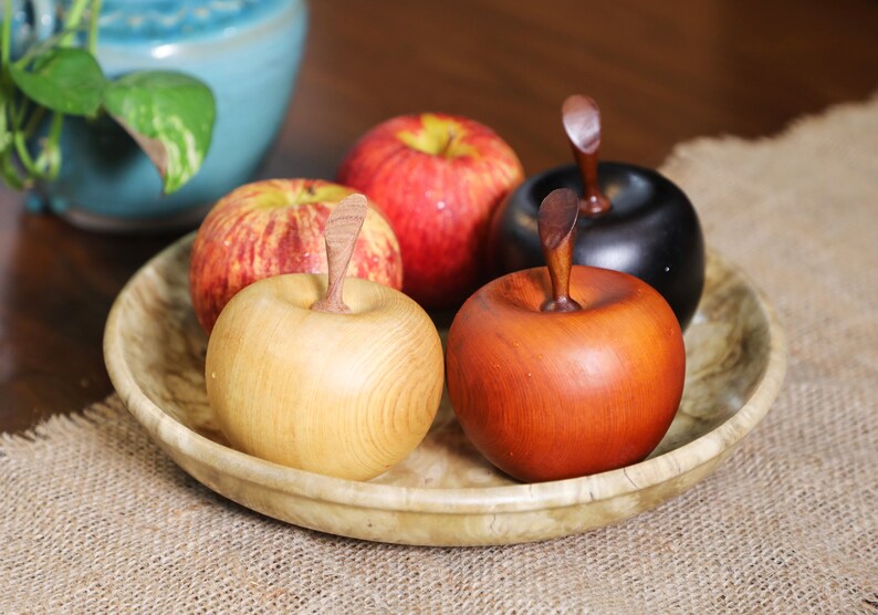 May include: Four wooden apples in a bowl. One apple is red, one is black, one is light brown, and one is dark brown.
