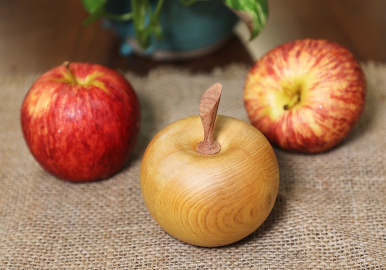 May include: Three apples, two red and one wooden, on a burlap surface. The wooden apple has a carved stem.