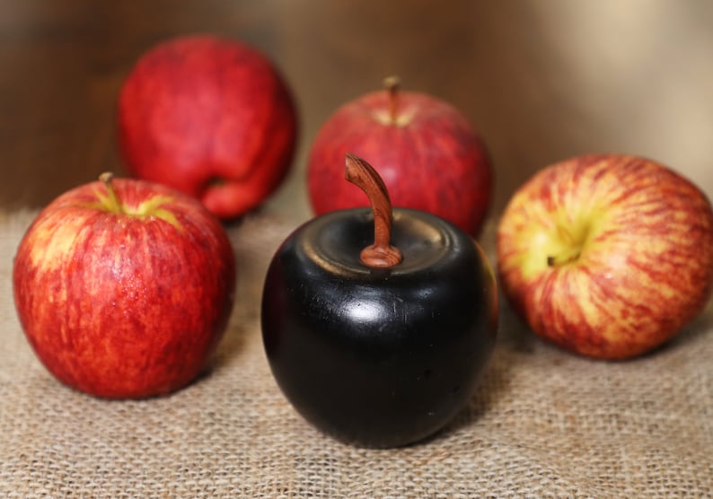 May include: Five apples, four red and one black, on a burlap surface. The black apple is made of wood and has a wooden stem.