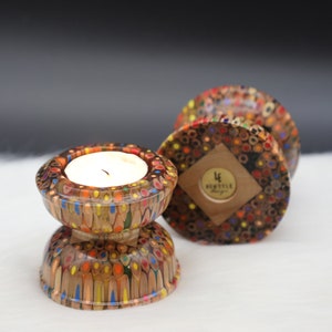 May include: Two candle holders made from colorful pencil shavings. The candle holders are in the shape of a bowl with a smaller bowl on top. The top bowl has a white candle inside. The bottom bowl has a diamond-shaped wooden label with the text "Lifestyle Designs".