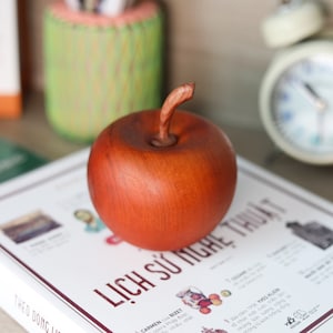 May include: A wooden apple sculpture with a reddish-brown finish. The apple is sitting on a white book with the text "LỊCH SỬ NGHỆ THUẬT" printed on the cover.