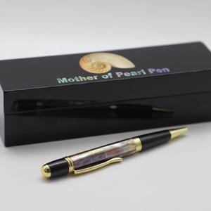 Puede incluir: Un bolígrafo negro con cuerpo de nácar y detalles dorados descansa junto a una caja negra. La caja tiene un acabado brillante y presenta un diseño de concha de nautilus y el texto "Mother of Pearl Pen".