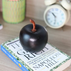 May include: A black apple figurine with a brown stem sits on top of a book titled "Think and Grow Rich" by Napoleon Hill. The book cover features a design of dollar bills.
