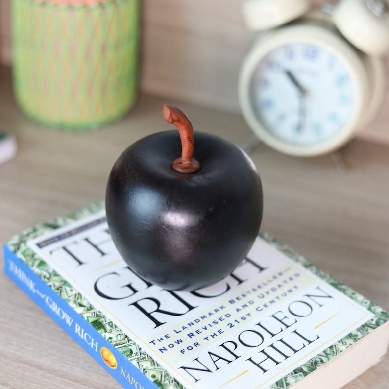 May include: A black apple figurine with a brown stem sits on top of a book titled "Think and Grow Rich" by Napoleon Hill. The book cover features a design of dollar bills.