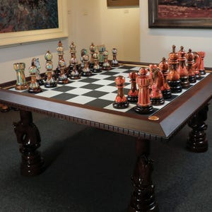 Puede incluir: Un juego de ajedrez ornamentado sobre una mesa de madera oscura. Las piezas del ajedrez son de varios colores, incluyendo rojo, dorado y negro. La mesa tiene un tablero de ajedrez y patas con forma de caballo. La mesa mide unos 150 cm de largo.