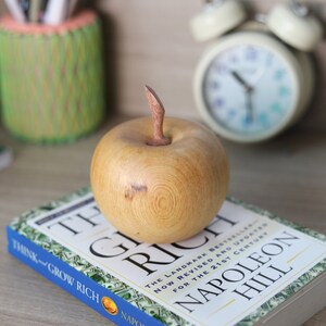 May include: A wooden apple sculpture with a light brown finish and a natural wood grain pattern. The apple is sitting on top of a book titled "Think and Grow Rich" by Napoleon Hill.