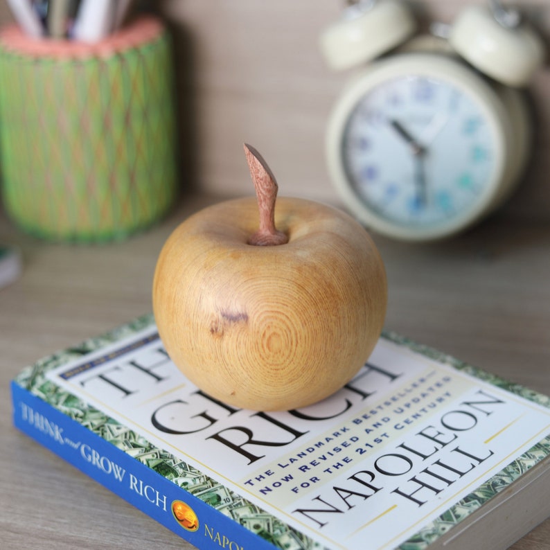 May include: A wooden apple sculpture with a light brown finish and a natural wood grain pattern. The apple is sitting on top of a book titled "Think and Grow Rich" by Napoleon Hill.