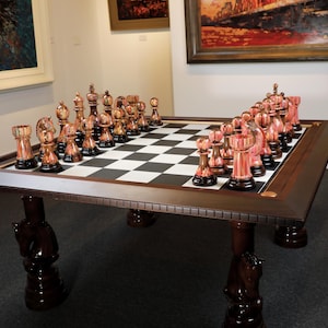 Puede incluir: Un juego de ajedrez ornamentado con una mesa de madera marrón oscuro y un tablero de juego a cuadros blanco y negro. Las piezas de ajedrez son de color cobre y están dispuestas para jugar. La mesa tiene patas decorativas.