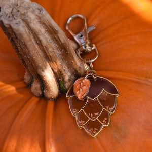May include: A close-up of a bright orange pumpkin with a brown stem. A gold-colored keychain with a brown and gold hop design is attached to the pumpkin. The keychain has a small orange leaf accent.