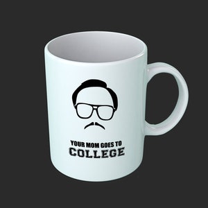 May include: White ceramic mug with a black graphic of a man with glasses and a mustache. The text "YOUR MOM GOES TO COLLEGE" is printed below the graphic. The mug has a curved handle and is set against a dark background.
