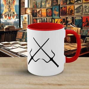 May include: A white ceramic mug with a bright red interior and a matching red handle. The exterior features a black graphic illustration of two crossed sai weapons centered on the front surface of the mug, creating a sharp contrast against the white.