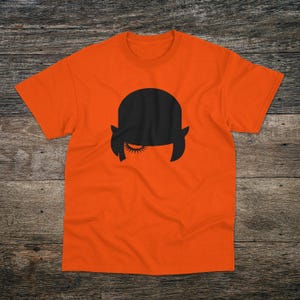 May include: Orange t-shirt with a black silhouette of a person wearing a bowler hat and a single eye open.