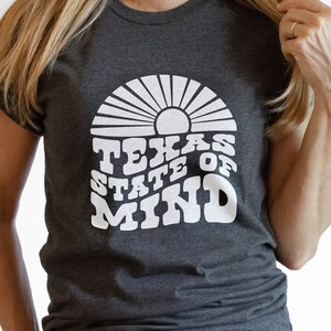 May include: A dark grey t-shirt with a white graphic design of a sun with rays and the text "Texas State of Mind".