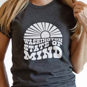 Può includere: Una maglietta grigia scura con una grafica bianca di un sole e il testo "Washington State of Mind".
