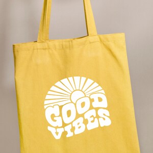 Peut inclure: Un sac fourre-tout en toile jaune avec du texte blanc qui dit "GOOD VIBES" dans une police r&eacute;tro. Le texte est entour&eacute; d'un motif de soleil blanc.