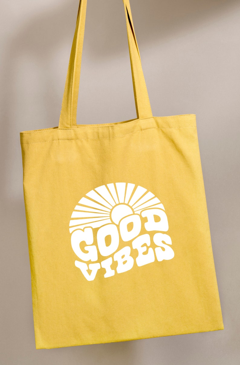 Peut inclure: Un sac fourre-tout en toile jaune avec du texte blanc qui dit "GOOD VIBES" dans une police r&eacute;tro. Le texte est entour&eacute; d'un motif de soleil blanc.