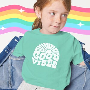 Peut inclure: Une jeune personne portant un t-shirt vert clair avec un motif blanc qui dit "GOOD VIBES" dans une police r&eacute;tro. Le motif comprend &eacute;galement un soleil avec des rayons. La personne porte une jupe blanche avec un lacet &agrave; l'avant et une veste en jean.