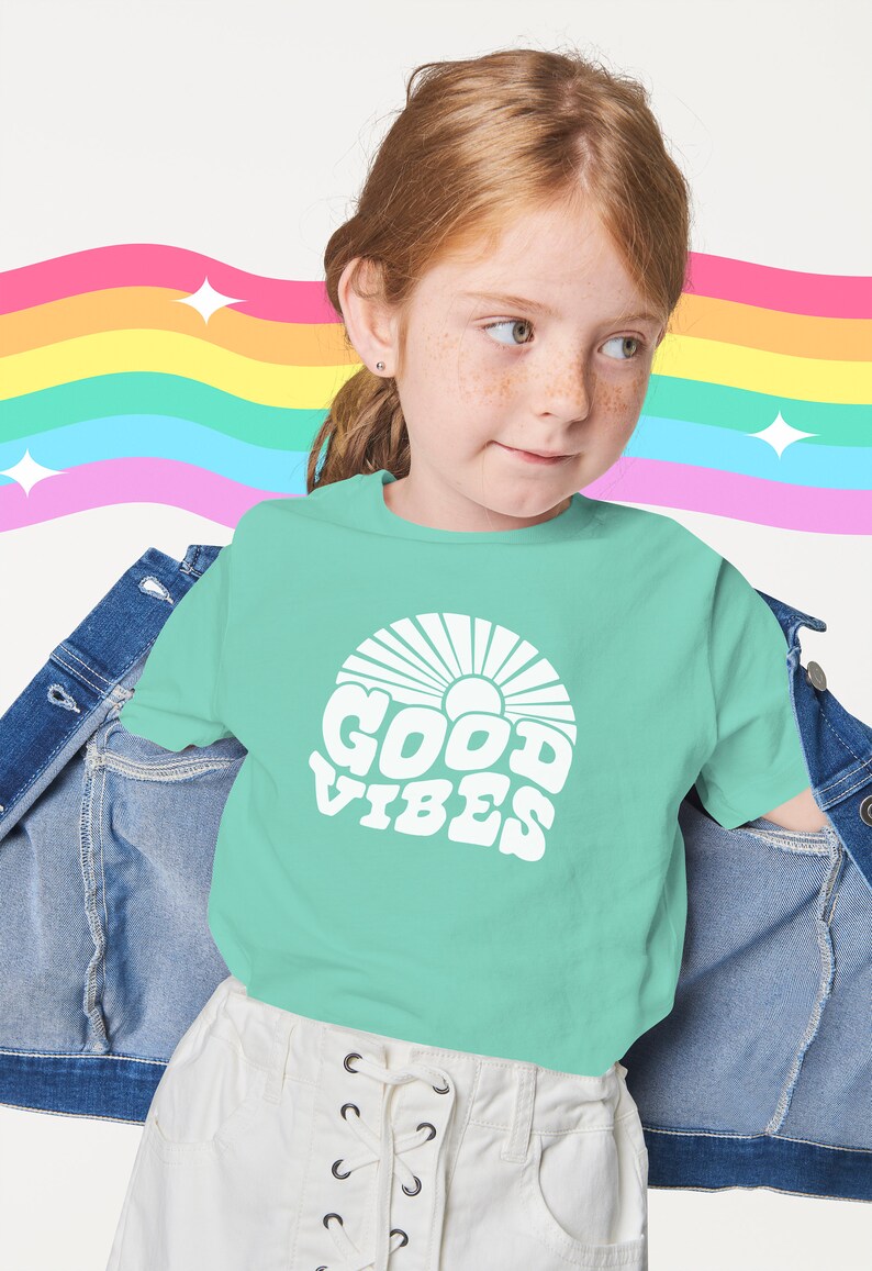 Peut inclure: Une jeune personne portant un t-shirt vert clair avec un motif blanc qui dit "GOOD VIBES" dans une police r&eacute;tro. Le motif comprend &eacute;galement un soleil avec des rayons. La personne porte une jupe blanche avec un lacet &agrave; l'avant et une veste en jean.