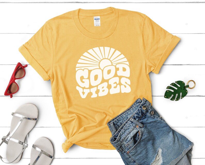 Peut inclure: Un t-shirt jaune avec un motif blanc repr&eacute;sentant un soleil et le texte "GOOD VIBES".