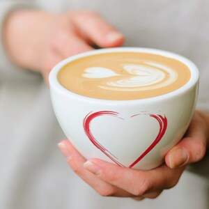 May include: A white ceramic mug with a red heart design, filled with a latte with a heart-shaped design in the foam.