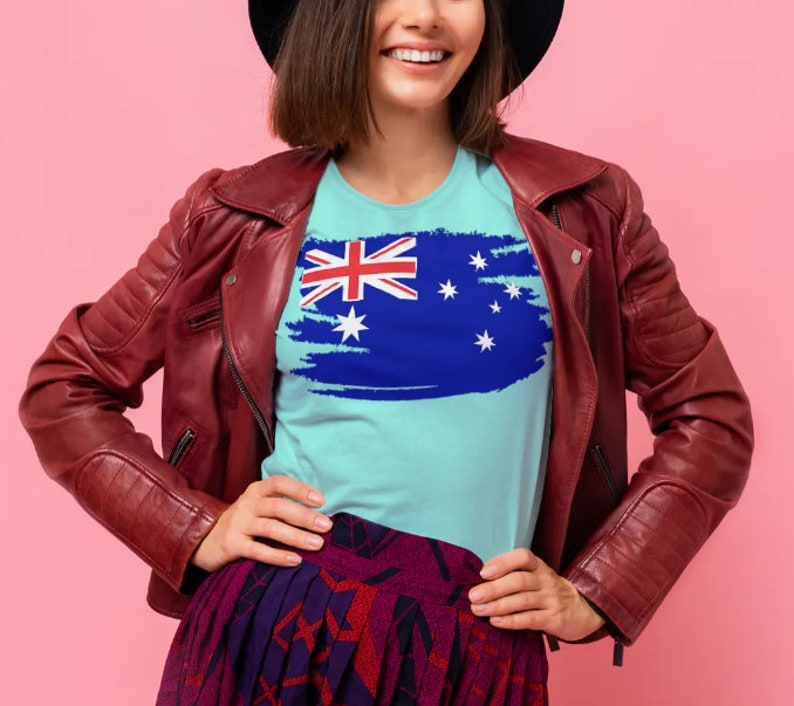 May include: A woman wearing a blue t-shirt with a distressed Australian flag design. She is wearing a red leather jacket and a black hat.
