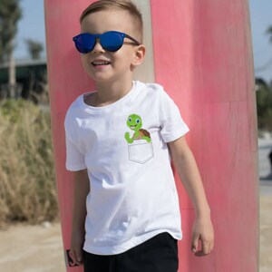 May include: A white t-shirt with a green turtle in the pocket. The turtle is smiling and has its arms raised. The shirt is being worn by a young boy wearing blue sunglasses.