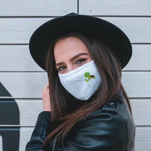 May include: A white face mask with a green turtle graphic in a pocket design. The mask is being worn by a person with long brown hair and a black hat.