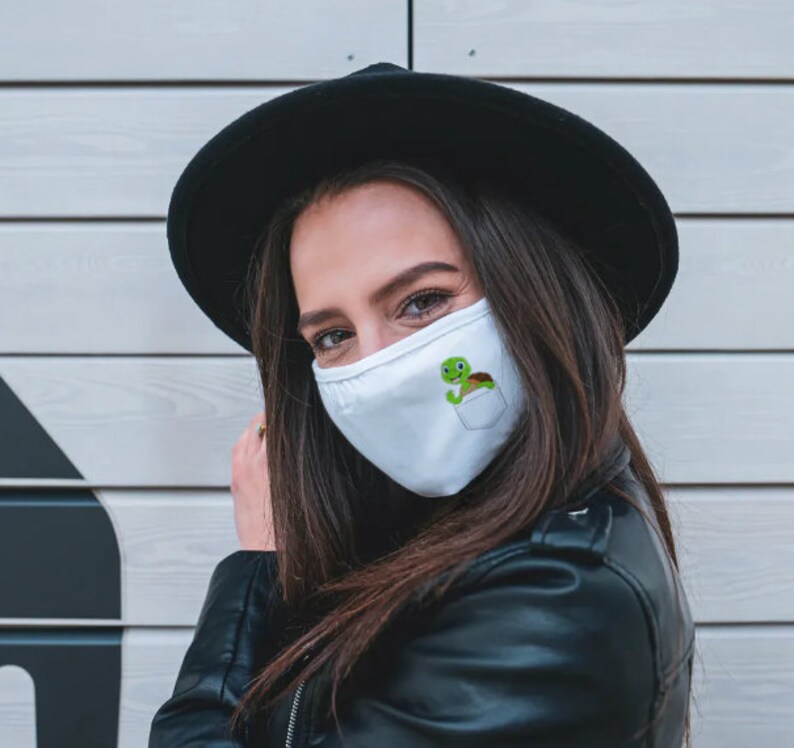 May include: A white face mask with a green turtle graphic in a pocket design. The mask is being worn by a person with long brown hair and a black hat.