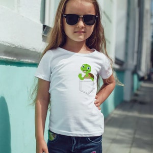 May include: A white t-shirt with a green turtle in a pocket. The turtle is smiling and has a brown shell. The girl wearing the shirt has long brown hair and is wearing sunglasses.