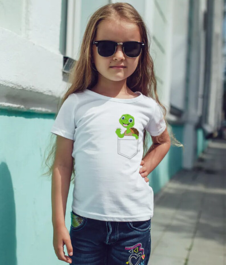 May include: A white t-shirt with a green turtle in a pocket. The turtle is smiling and has a brown shell. The girl wearing the shirt has long brown hair and is wearing sunglasses.