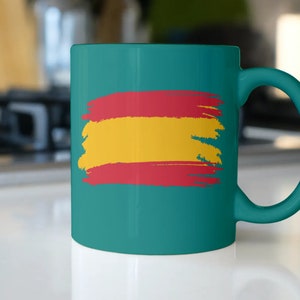 May include: A teal ceramic mug with a painted Spanish flag design. The flag is red, yellow, and red with a brushstroke effect.