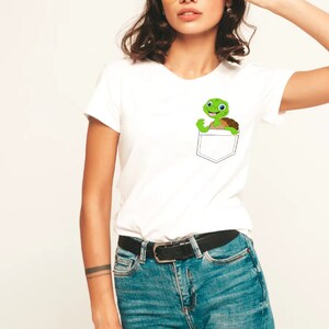 May include: White t-shirt with a small pocket on the left side. A cartoon turtle is peeking out of the pocket.