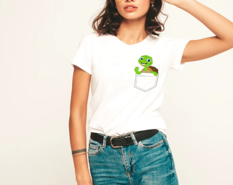 May include: White t-shirt with a small pocket on the left side. A cartoon turtle is peeking out of the pocket.