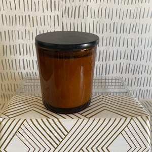 May include: A brown glass candle jar with a black lid. The jar is cylindrical and sits on a white and gold geometric patterned surface. The background features a white and gray vertical line pattern.