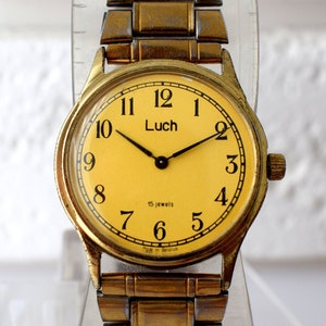 May include: A vintage-style wristwatch with a round gold-tone case and a matching metallic link bracelet. The watch face is a solid pale yellow color featuring black Arabic numerals, black hands, and the brand name Luch printed in black.