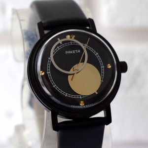 May include: A black wristwatch with a black leather band and a round face. The face has a gold circle in the center and the word "RAKET" printed on it.