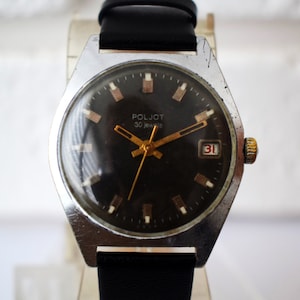 Puede incluir: Un reloj de pulsera plateado con una esfera negra y agujas doradas. El reloj tiene una ventana de fecha en la posición de las 3 en punto y dice "POLJOT 30 jewels" en la esfera.