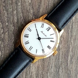 May include: A gold-toned Raketa wristwatch with a white face and black Roman numerals. The watch has a black leather band. The watch face displays the Raketa brand name. The watch is on a wooden surface.