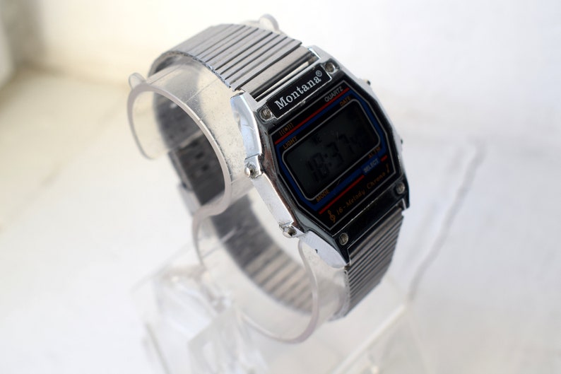 K&ouml;nnte beinhalten: Eine silberfarbene Montana Digitaluhr mit schwarzem Zifferblatt, das die Uhrzeit anzeigt. Die Uhr hat ein rechteckiges Geh&auml;use mit blauen und roten Akzenten. Das Armband besteht aus silberfarbenen Metallgliedern.