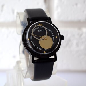 May include: A black wristwatch with a black leather band and a gold face. The face has a black circle in the center with a gold circle around it. The watch says "PAKETA" on the face.