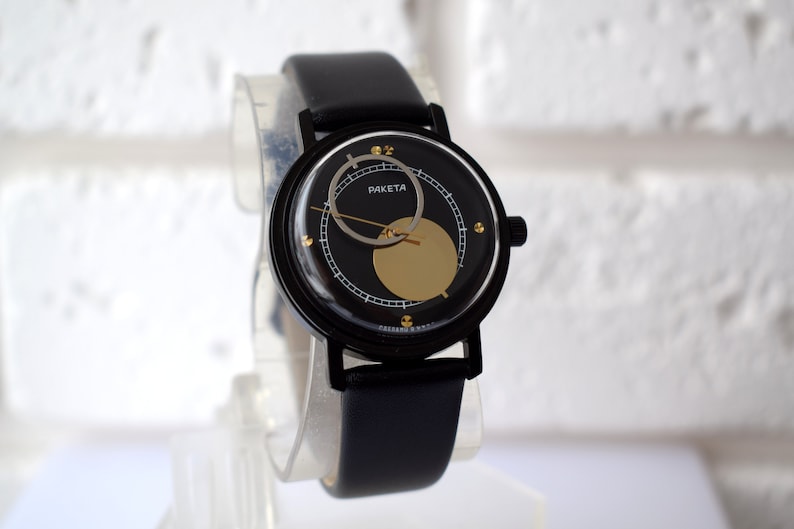 May include: A black wristwatch with a black leather band and a gold face. The face has a black circle in the center with a gold circle around it. The watch says "PAKETA" on the face.