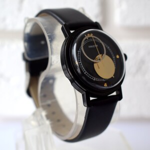 May include: A black wristwatch with a black leather band and a circular face. The face has a gold-colored circle in the center and a smaller circle around it. The watch face also has black markings and the word "RAKET" printed on it.